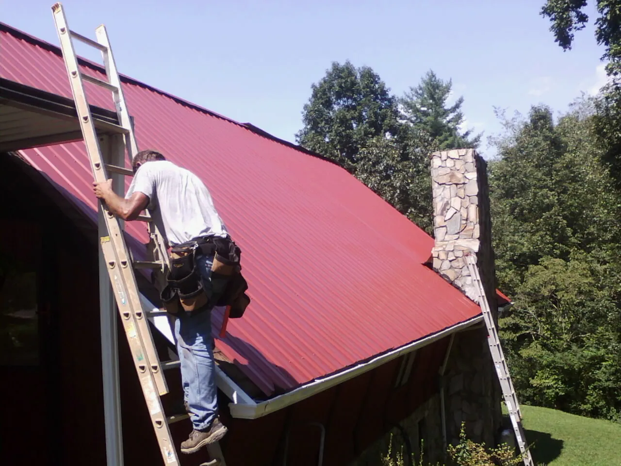 Roofer on a ladder beside a red metal roof and stone chimney — Soffit & Fascia Repair by Roofing Brothers in Carnegie